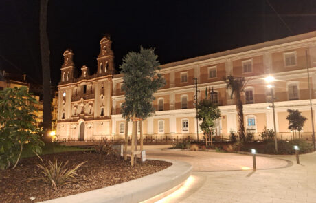 Plaza con mobiliario urbano de hormigón iluminado y un edificio histórico.