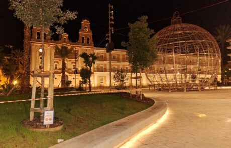 Plaza iluminada con bancos de hormigón prefabricado, esfera decorativa y edificio histórico al fondo.