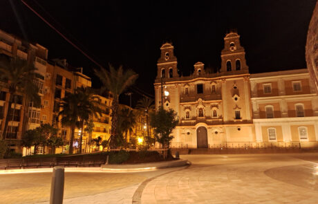 Plaza nocturna con bancos de mobiliario urbano de hormigón prefabricado frente a iglesia iluminada.