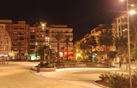 Plaza urbana iluminada de noche con bancos curvos de hormigón prefabricado y zonas ajardinadas.