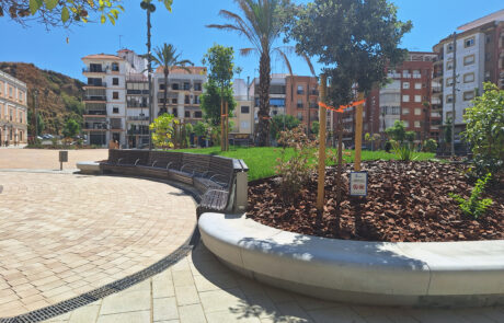 Plaza con banco curvado de hormigón, áreas verdes y edificios residenciales en un día soleado.