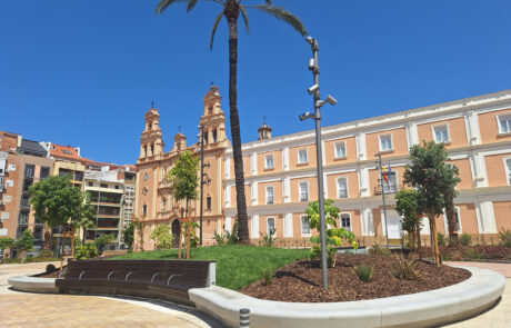 Plaza soleada con banco curvado de hormigón, palmera y edificio histórico al fondo.