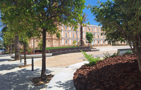 Plaza soleada con elementos urbanos de hormigón, bancos, árboles y edificios históricos.