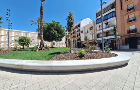 Banco curvo de hormigón prefabricado en una plaza soleada con jardines y edificios.