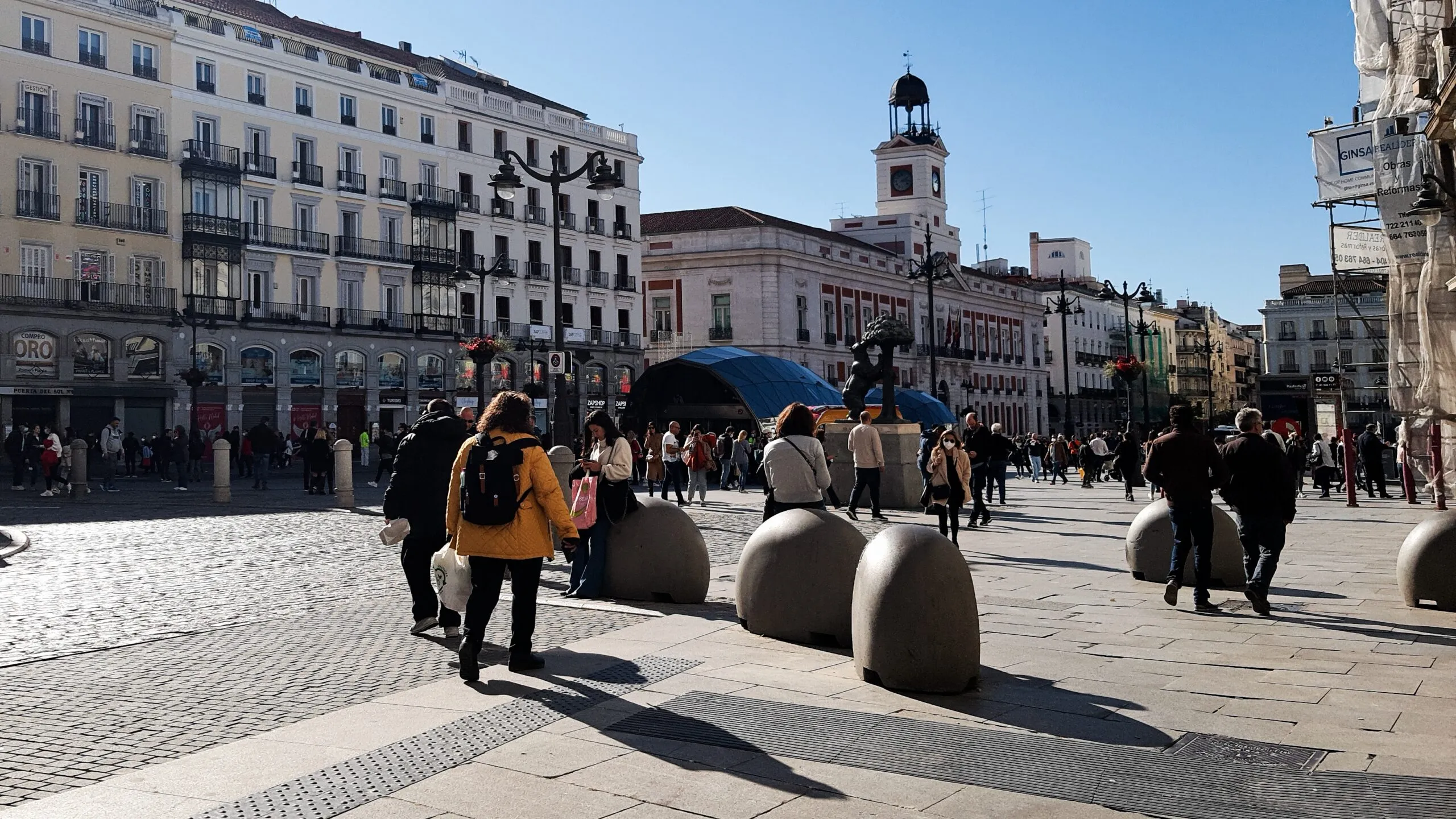 Personas caminando en una plaza con diversos elementos urbanos en forma de bolardos de hormigón frente a un edificio.